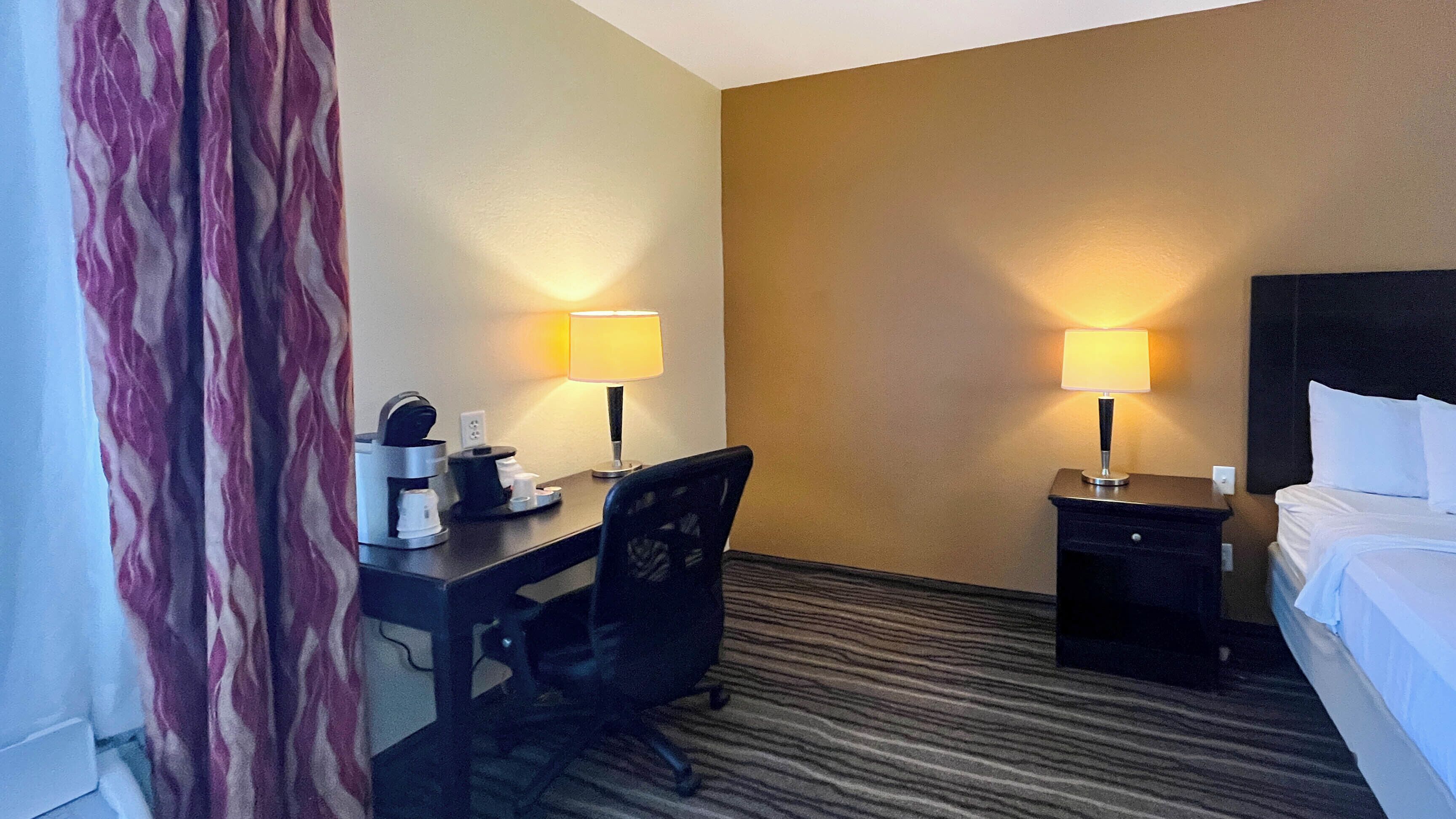 Quarto de hotel com uma cama, duas mesas de cabeceira com luminárias, uma escrivaninha com cafeteira e uma cadeira.