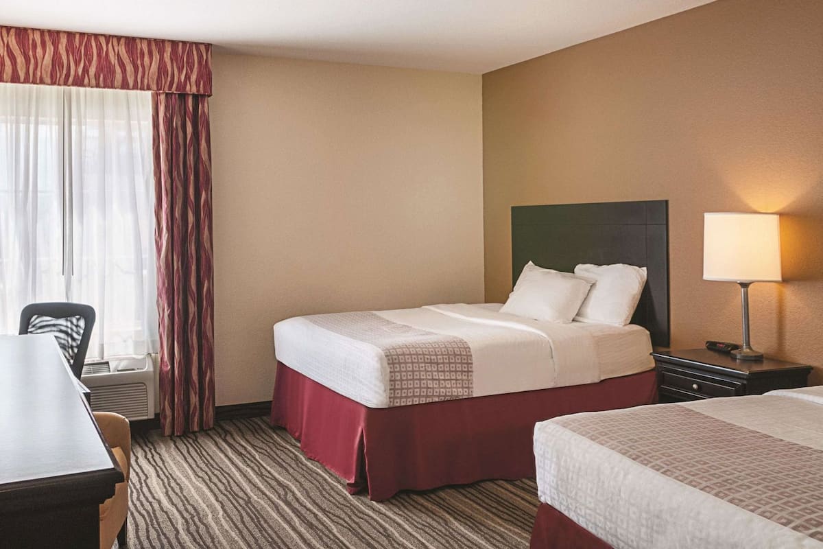 A hotel room with two beds, a desk, a chair, a lamp, and a window with curtains.