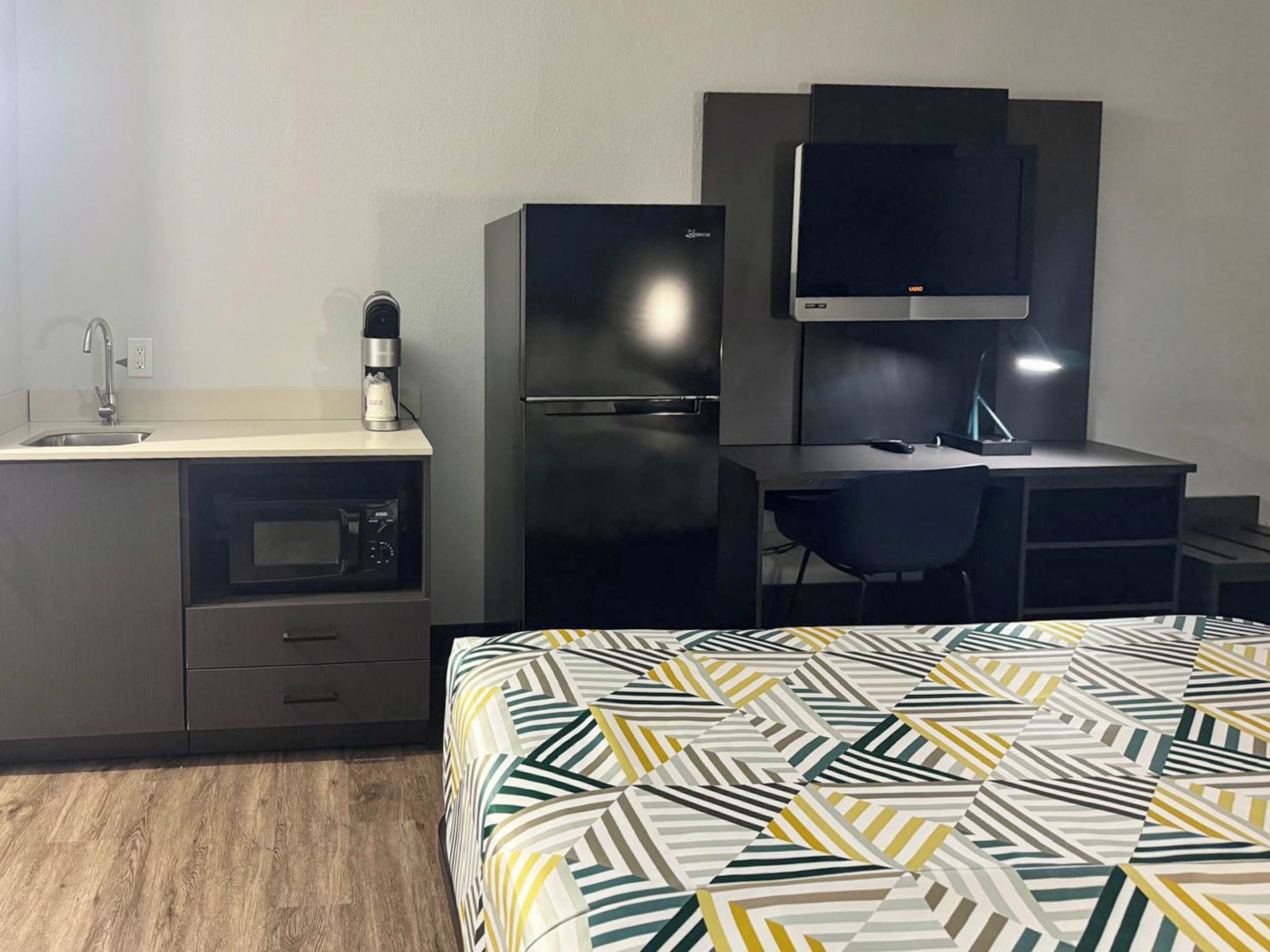 Habitación de hotel con cama, escritorio con silla, microondas, refrigerador y lavabo.