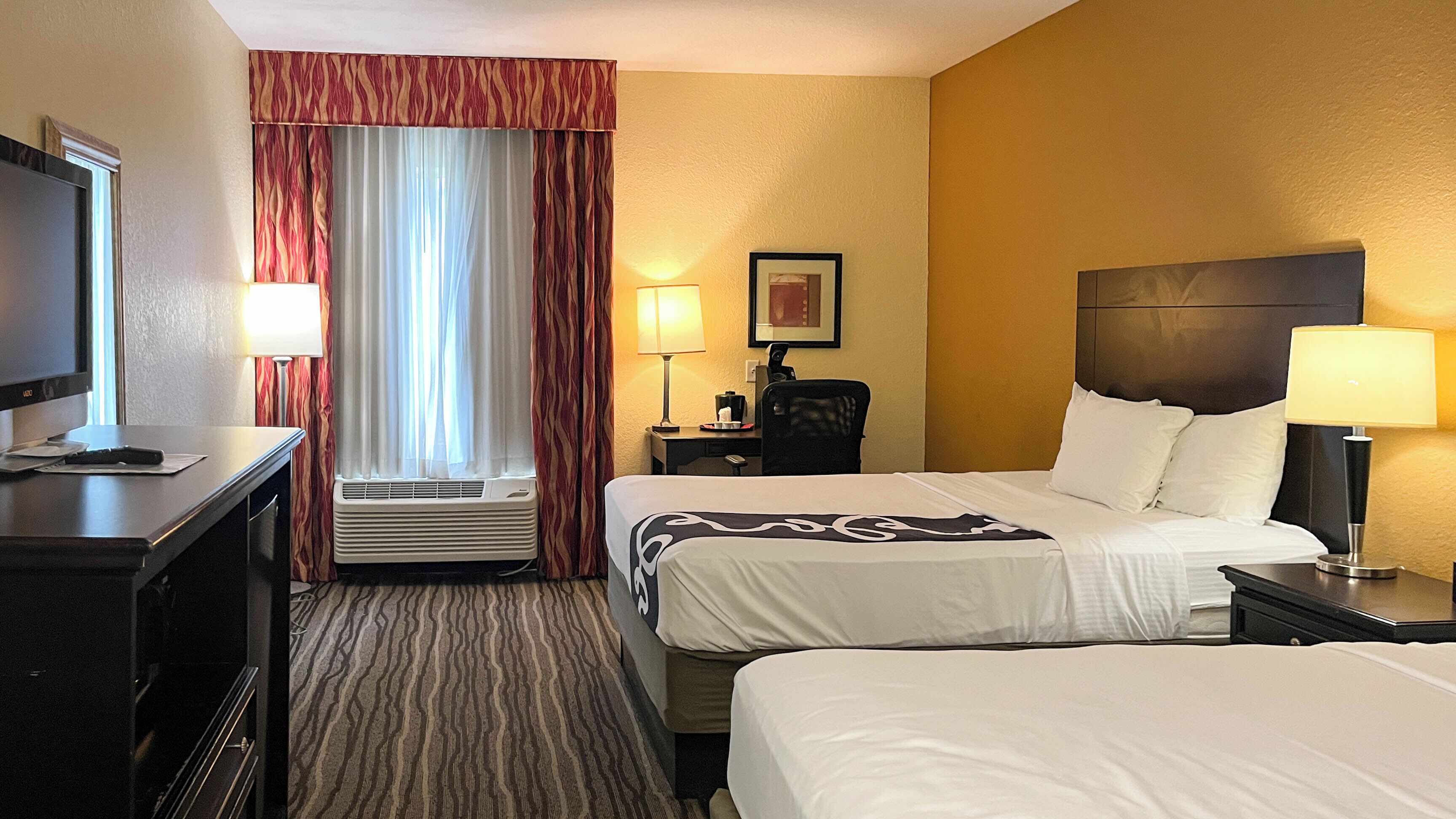 A hotel room with two beds, a desk with a TV, a desk lamp, a chair, a window with curtains, an air conditioner, and a framed picture on the wall.