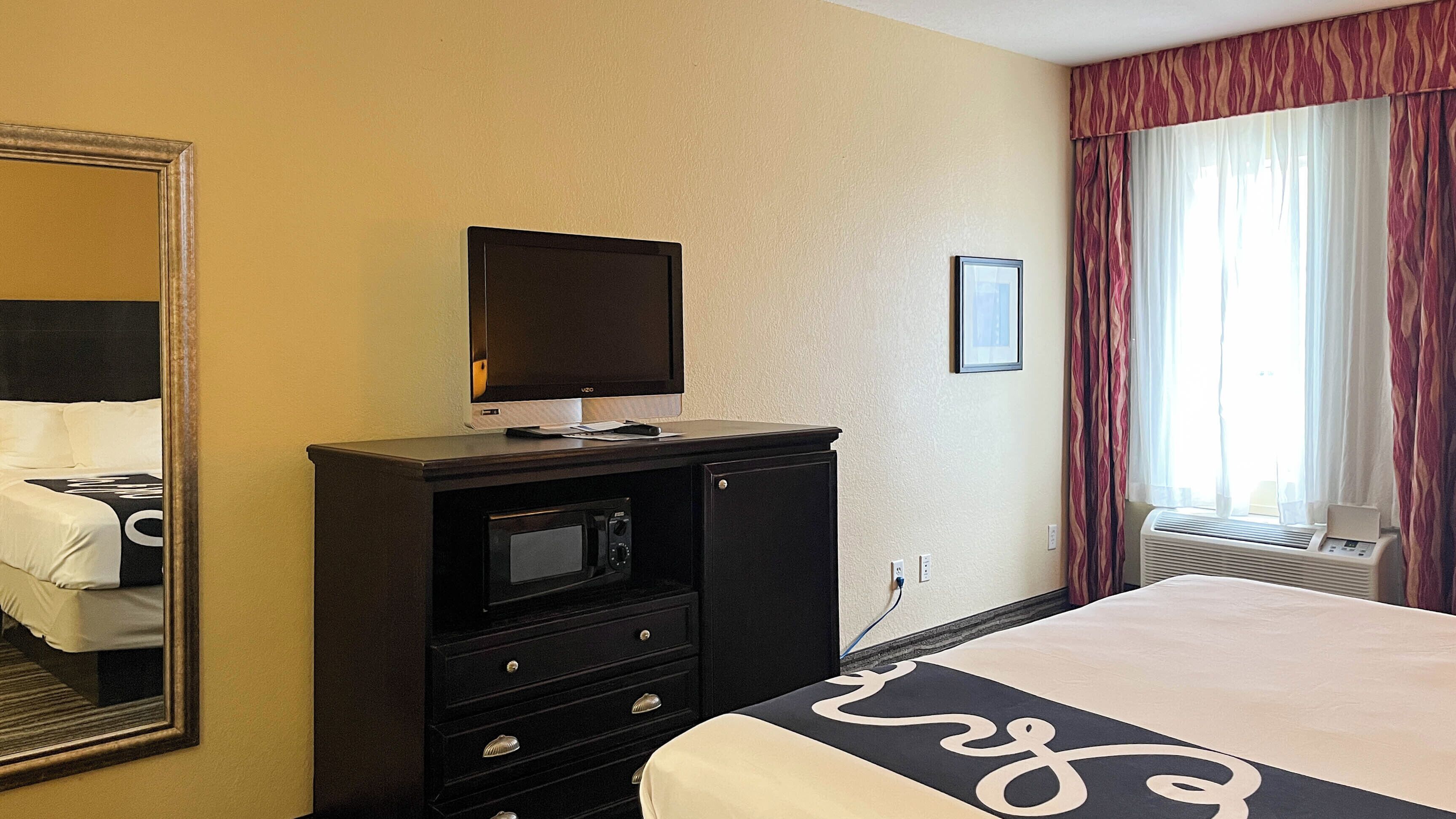 A hotel room with a bed, a TV on a dresser, a mirror, a window with curtains, and an air conditioning unit.