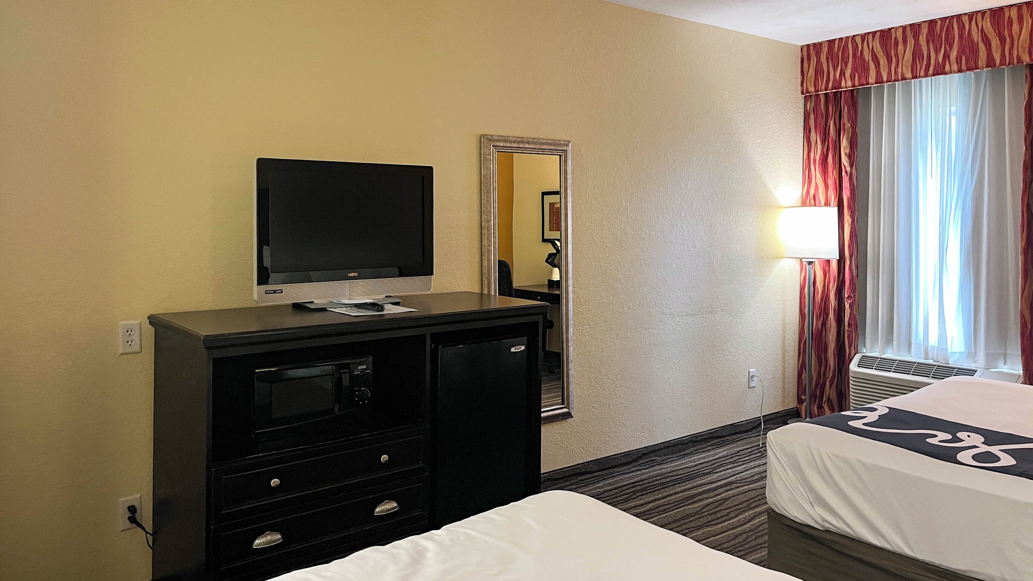 A hotel room with a TV, a desk with a lamp, a mirror, and a bed.