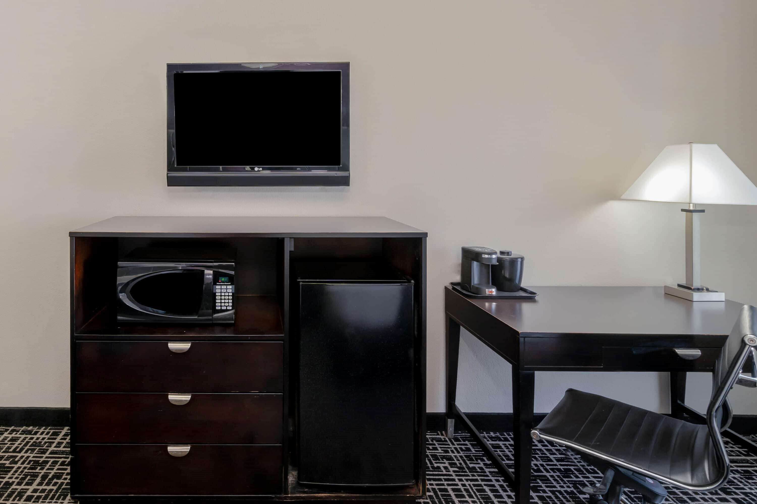 A hotel room with a flat-screen TV mounted on the wall, a minibar with a microwave, a coffee maker, and a desk with a chair.