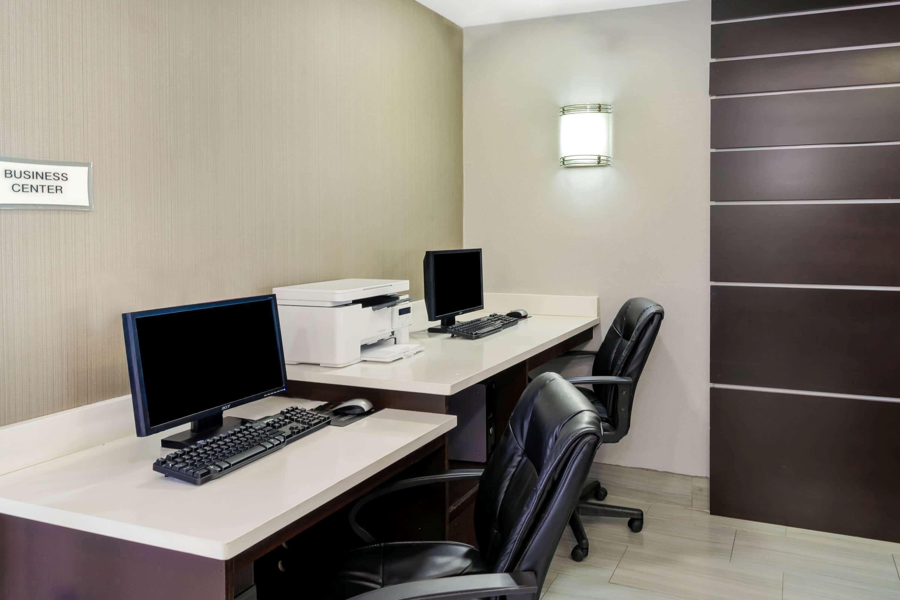 A business center with two desks, each equipped with a computer monitor, keyboard, and printer. There are black office chairs and a wall-mounted light fixture.