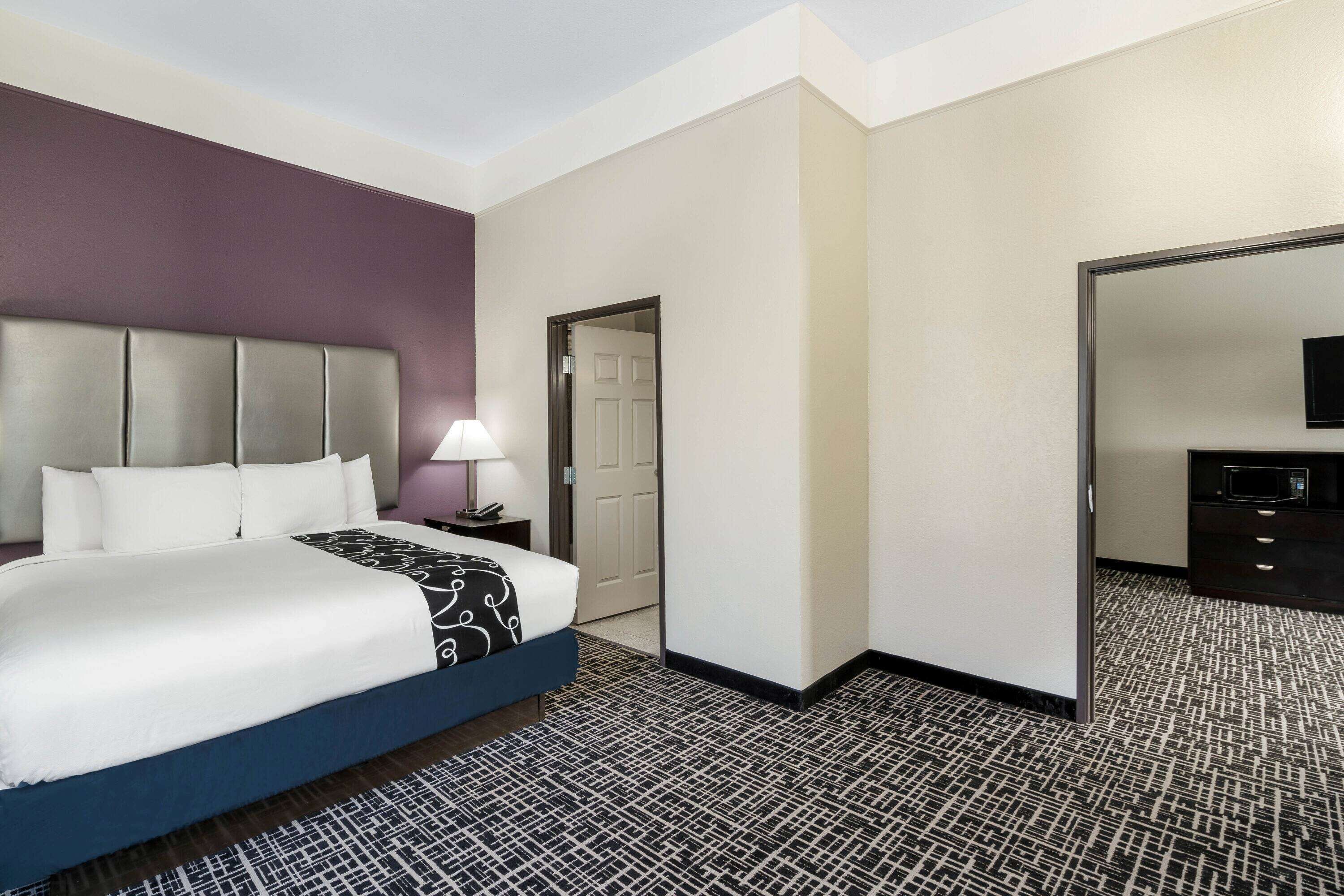 A hotel room with a bed, a nightstand, a lamp, a door, and a dresser.