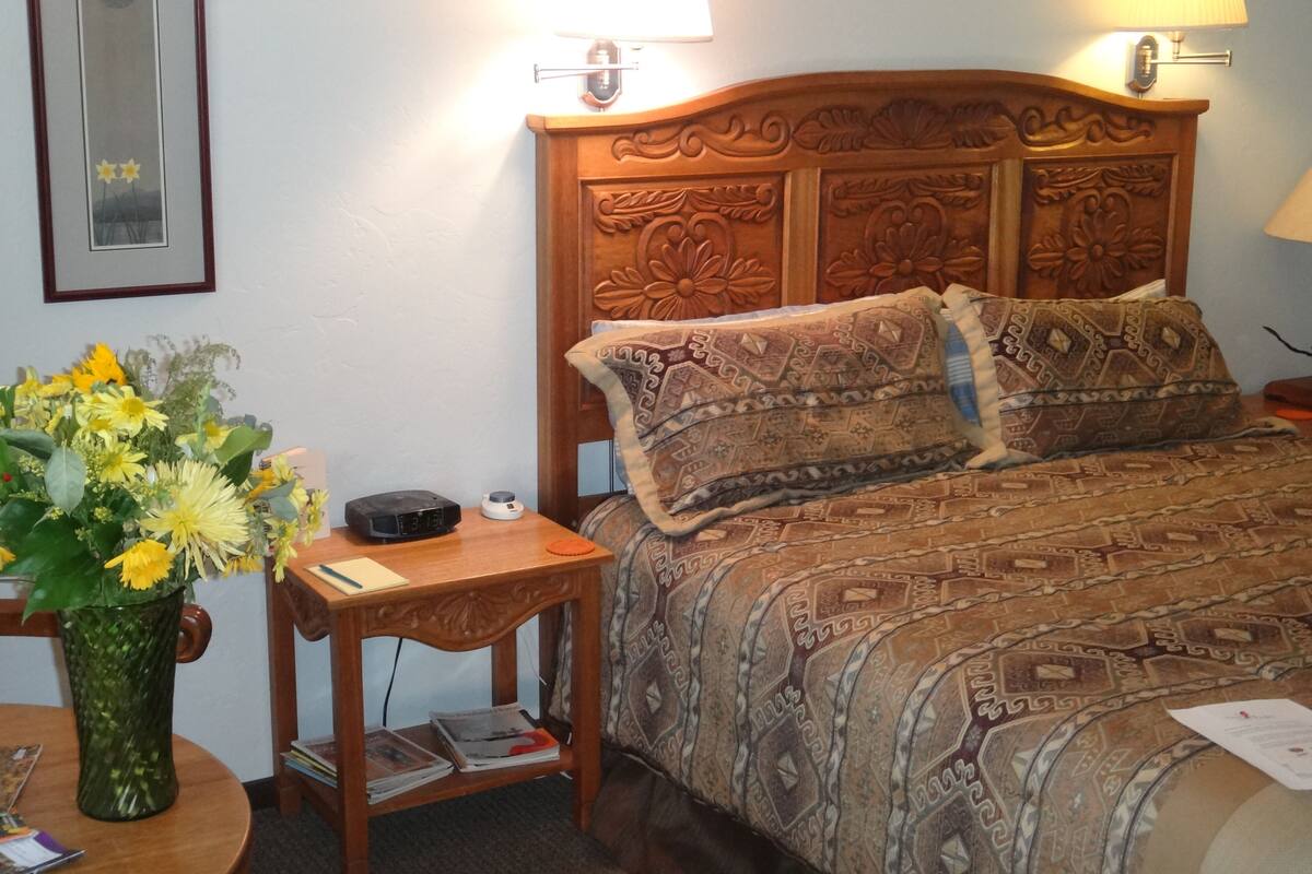 Quarto de hotel com cabeceira de madeira, mesa de cabeceira com abajur, um vaso de flores e cama com roupa de cama estampada.