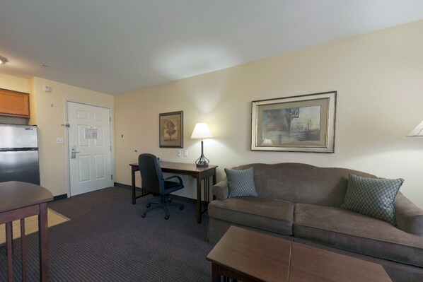 A hotel room with a kitchenette, a desk with a chair, a sofa, and a lamp.