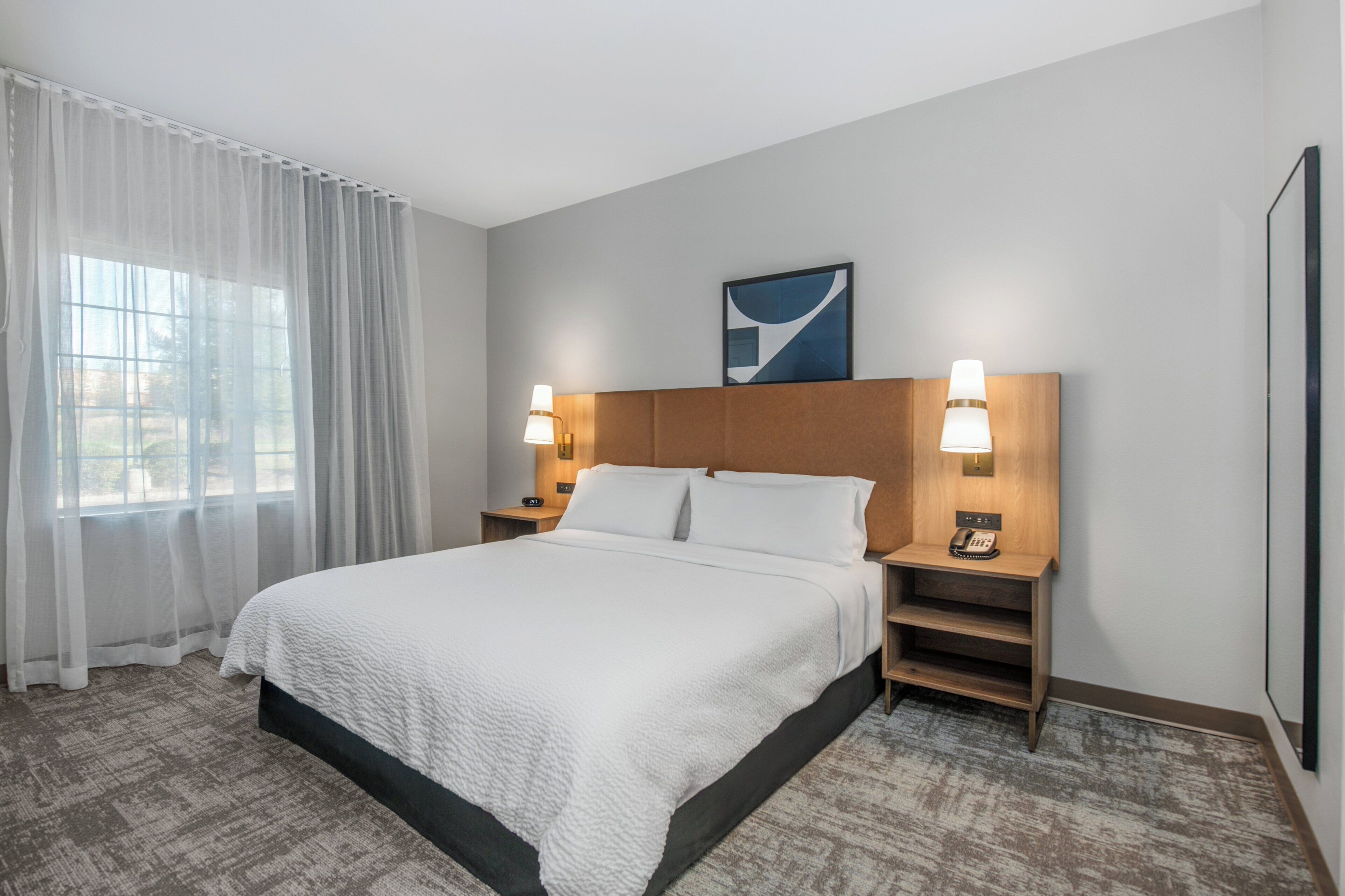 A hotel room with a large bed, two bedside tables with lamps, a headboard, and a window with sheer curtains.