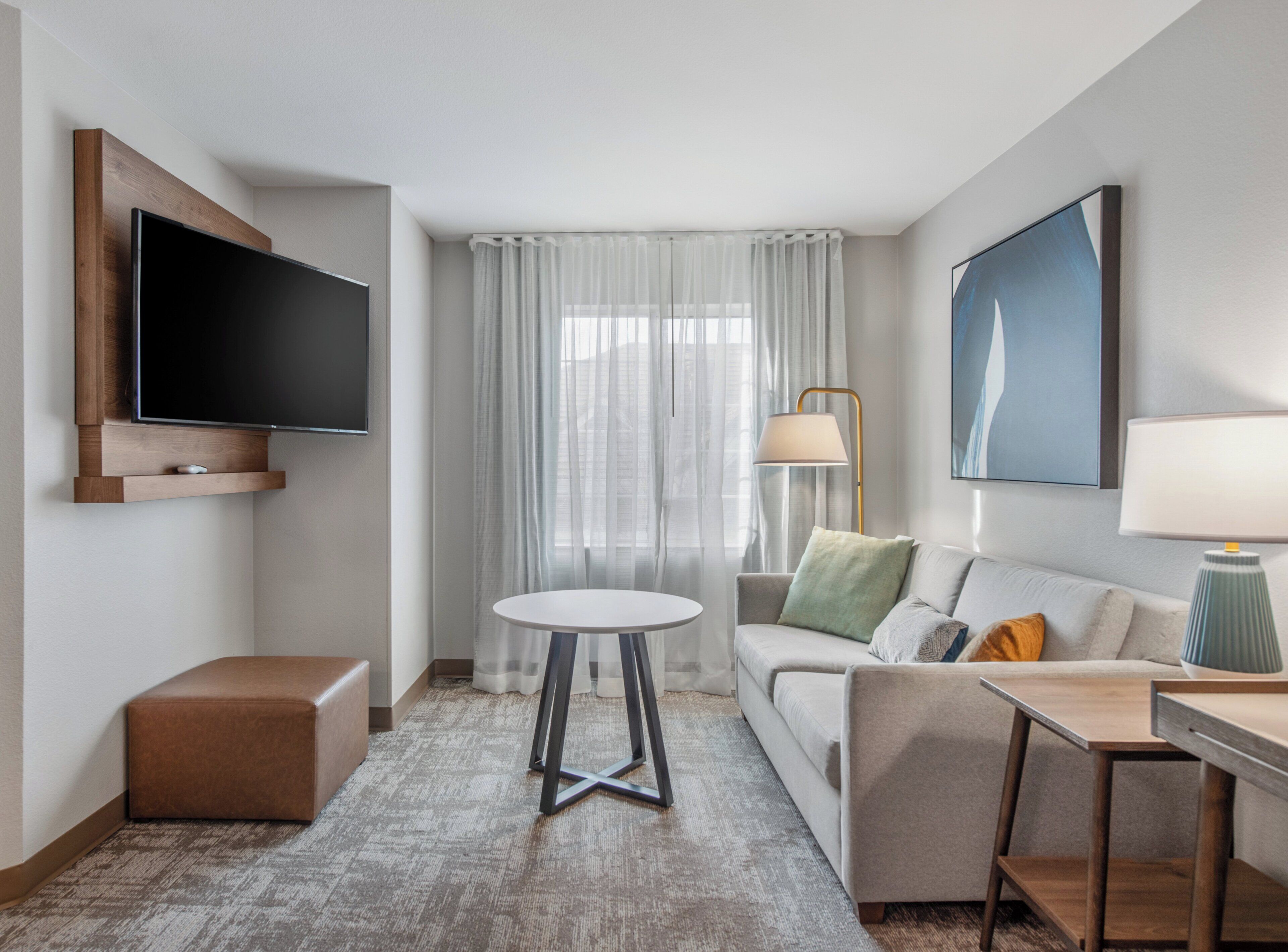 A modern hotel room with a flat-screen TV mounted on the wall, a round table, a sofa with cushions, a desk with a lamp, and a large window with sheer curtains.