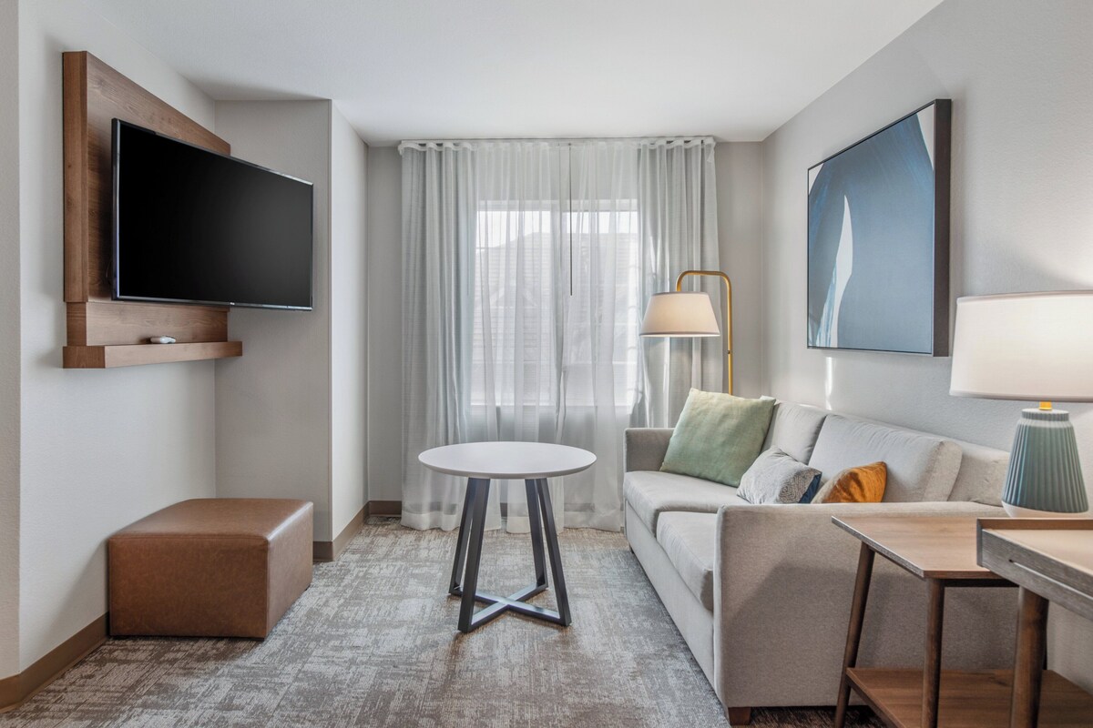 Ein modernes Hotelzimmer mit flachbildfernseher an der Wand, einem runden Tisch, einer Couch mit Kissen, einem Schreibtisch mit Lampe und einem großen Fenster mit durchsichtigen Vorhängen.