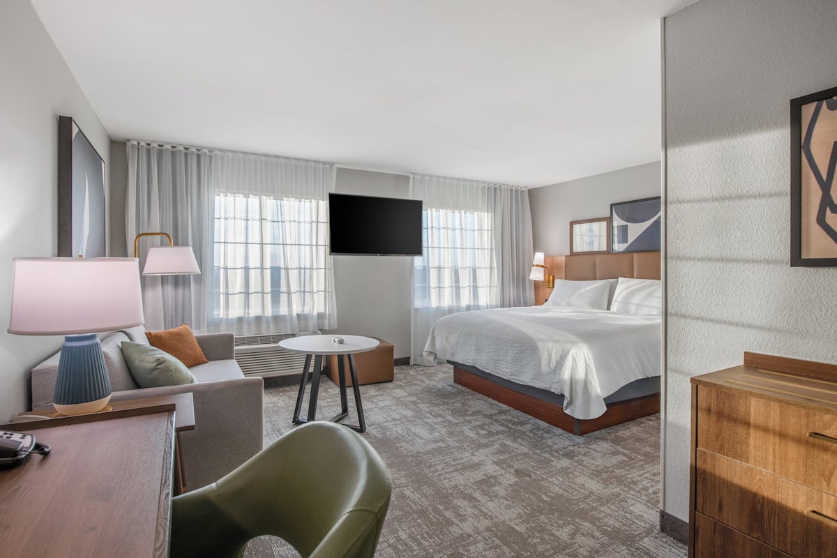A hotel room with a bed, a desk with a lamp, a chair, a TV, and a window with curtains.