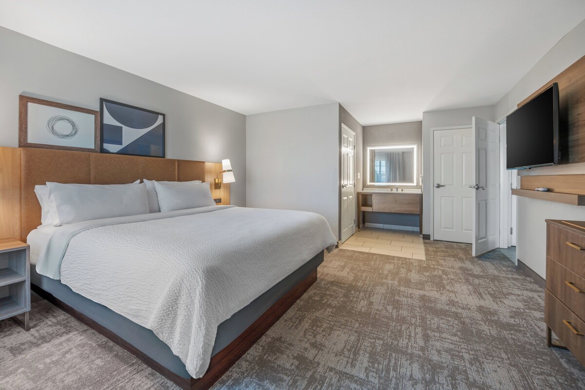 Ein modernes Hotelzimmer mit einem großen Bett, einem Fernseher, einem Schreibtisch und einem durch eine geöffnete Tür einsehbarer Badezimmer.