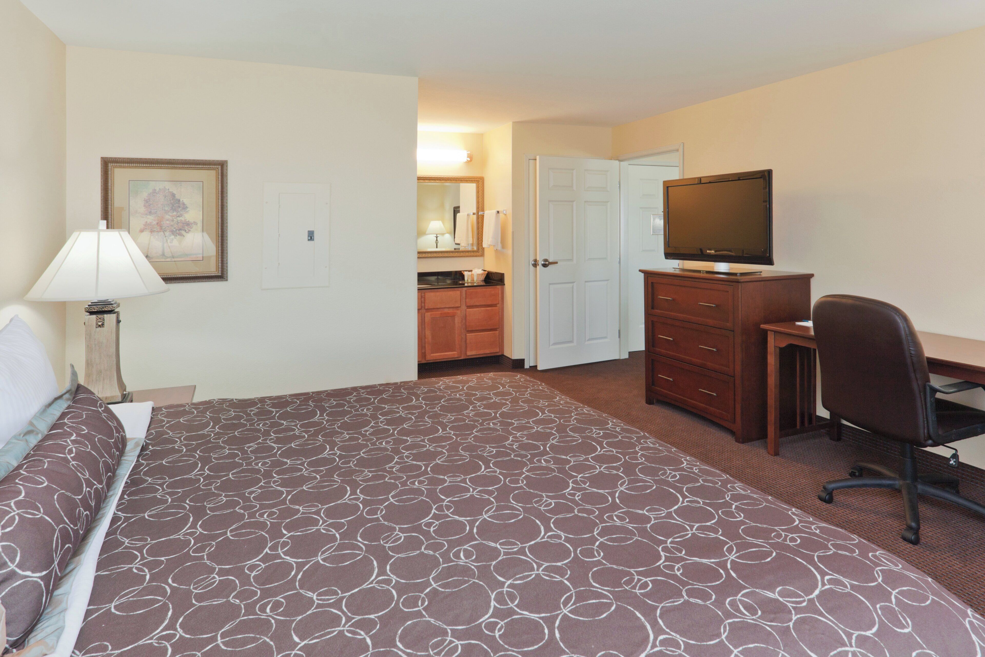 Une chambre d’hôtel avec un lit, un bureau, une chaise, une télévision, une commode et une salle de bain visible à travers une porte ouverte.