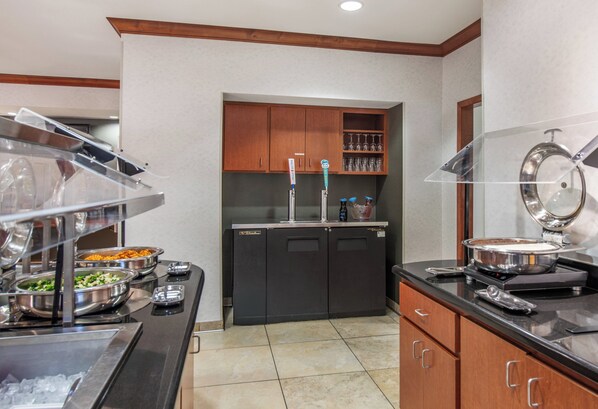 Un área de bufé con distintas estaciones de comida, una máquina de café y un refrigerador.