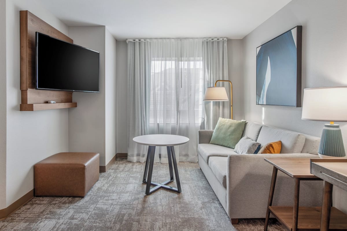 A modern hotel room with a flat-screen TV mounted on the wall, a round table, a sofa with cushions, a desk with a lamp, and a large window with sheer curtains.