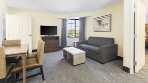 A hotel room with a dining table, chairs, a sofa, a TV, a window with curtains, and a framed picture on the wall.