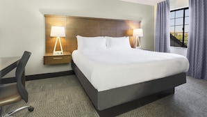 A hotel room with a large bed, two bedside lamps, a desk with a chair, and a view of the outdoors through a window.