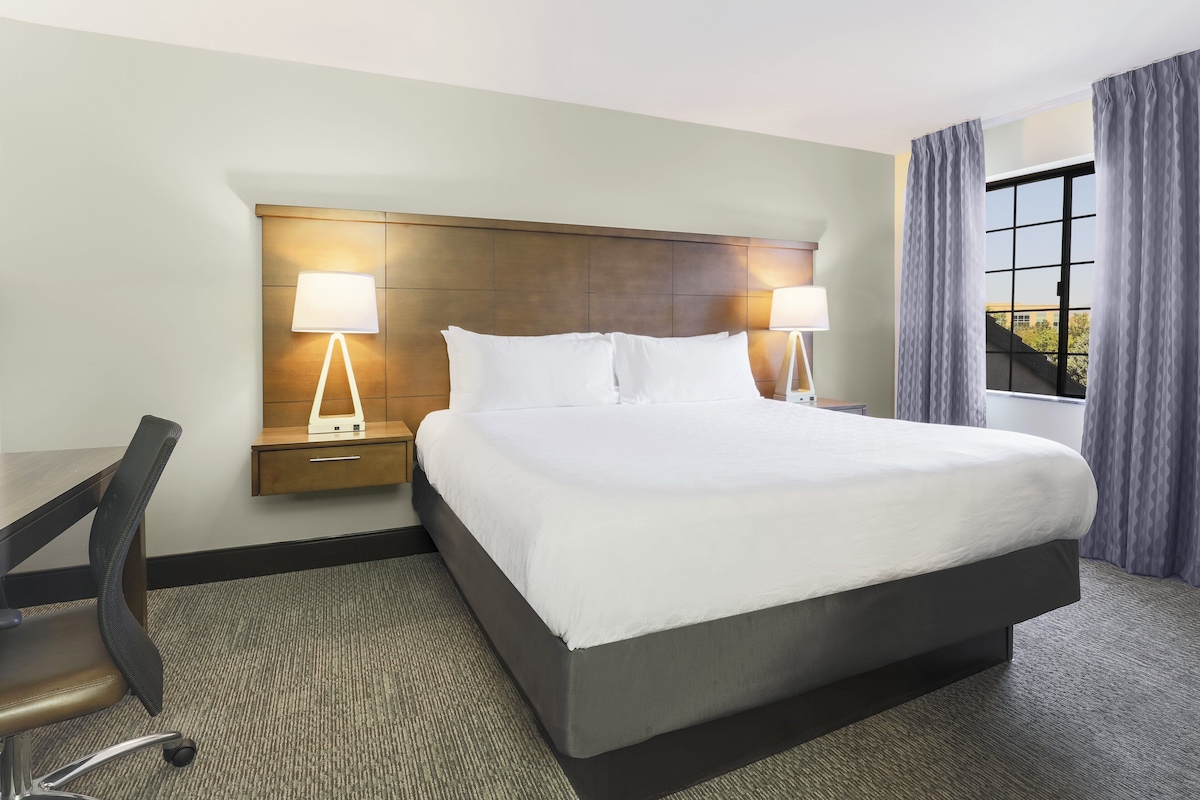 A hotel room with a large bed, two bedside lamps, a desk with a chair, and a view of the outdoors through a window.