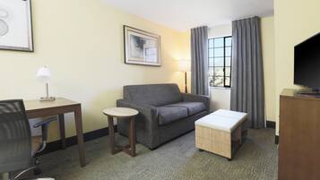 A hotel room with a sofa, a small table, a desk, a chair, a TV, and a window with curtains.