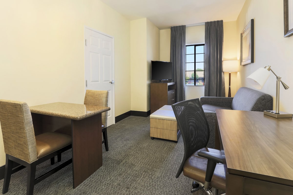 Ein Hotelzimmer mit Schreibtisch, zwei Stühlen, einer Couch, einem Fernseher, einer Lampe und einem Fenster mit Vorhängen.