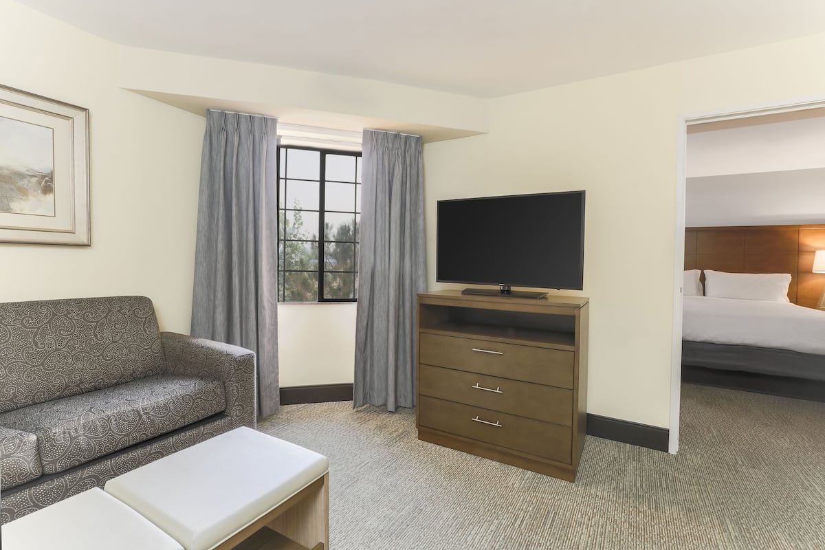 Ein Hotelzimmer mit einer Couch, einem Fernseher auf einem Holzboden, einem Bett und einem Fenster mit Vorhängen.