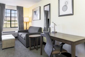 A hotel room with a sofa, a small table, a desk, a chair, a lamp, and a window with curtains.