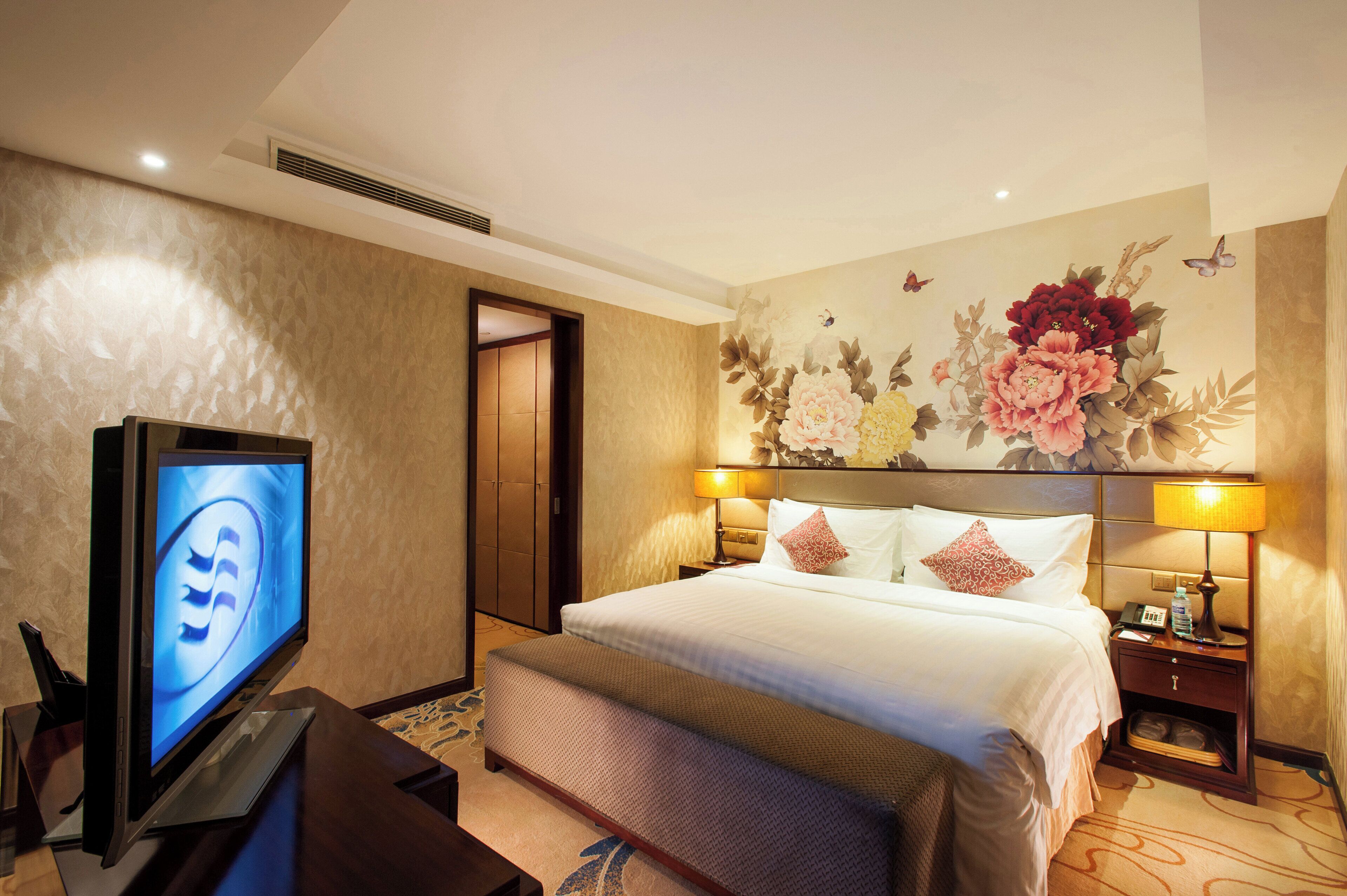 Une chambre d’hôtel équipée d’un lit, de lampes de chevet, d’une télévision, d’un bureau avec un ordinateur et d’une décoration murale représentant des motifs floraux.