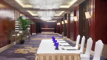 Un salón de conferencias de hotel con una larga mesa dispuesta para una reunión, rodeada de sillas blancas, una araña y un suelo alfombrado.