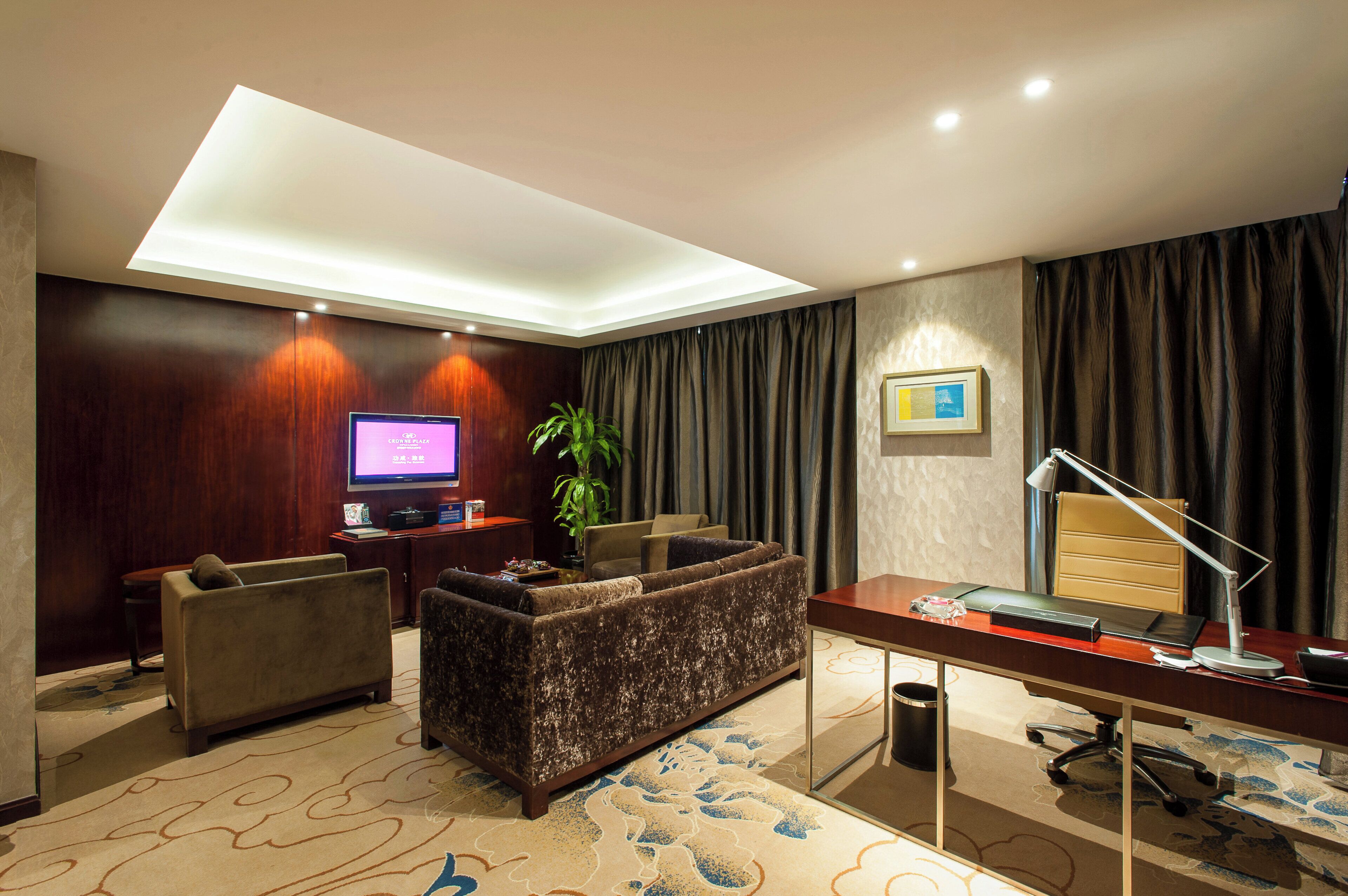 A hotel room with a sofa, a desk, a television, a plant, and a large window with curtains.