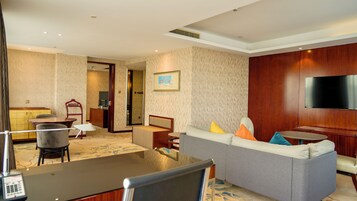 A hotel room with a living area, a dining table, a sofa with cushions, a TV mounted on the wall, and a desk with a chair.