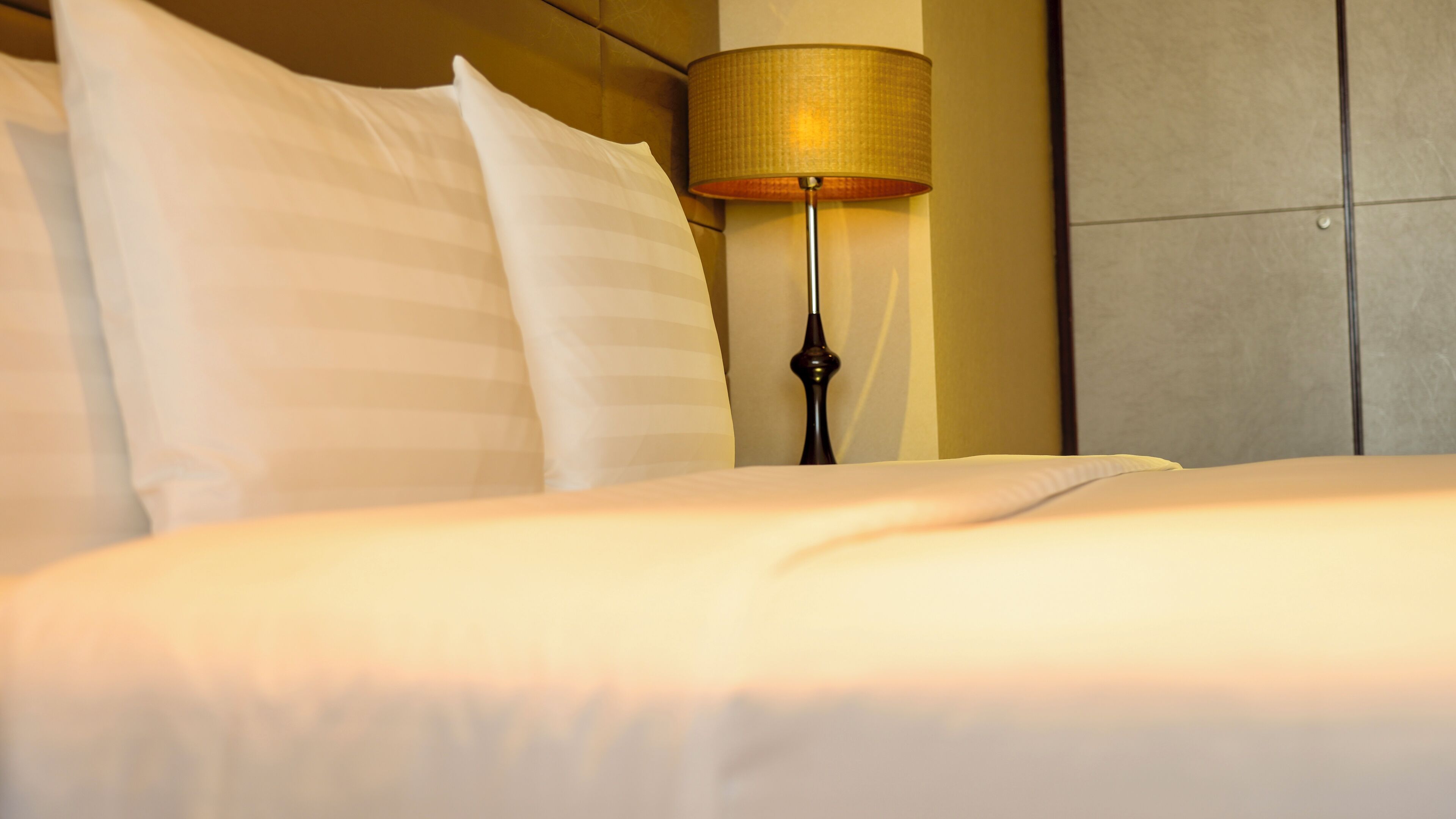 Una cama bien hecha con sábanas blancas, una lámpara de noche con pantalla color beige y un cabecero frente a una pared con textura.