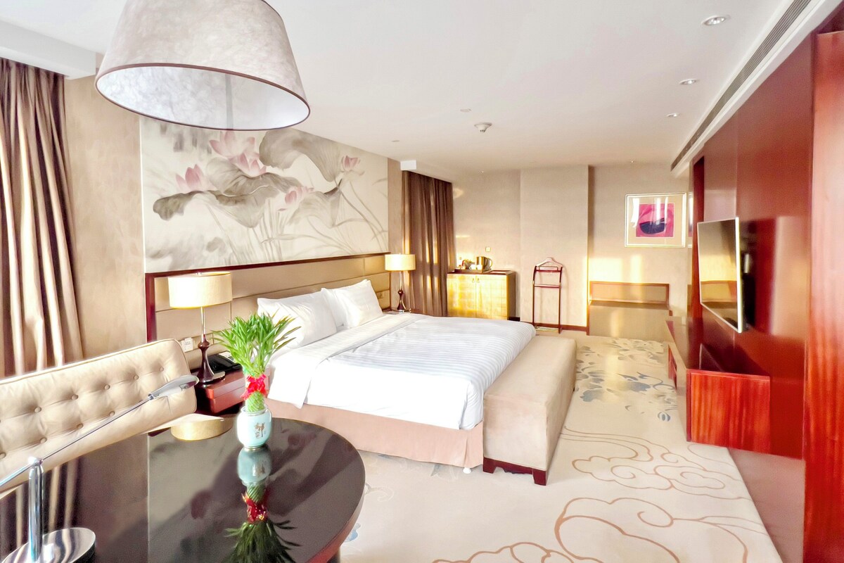 A modern hotel room with a large bed, a dining table, a TV, and a decorative wall piece.