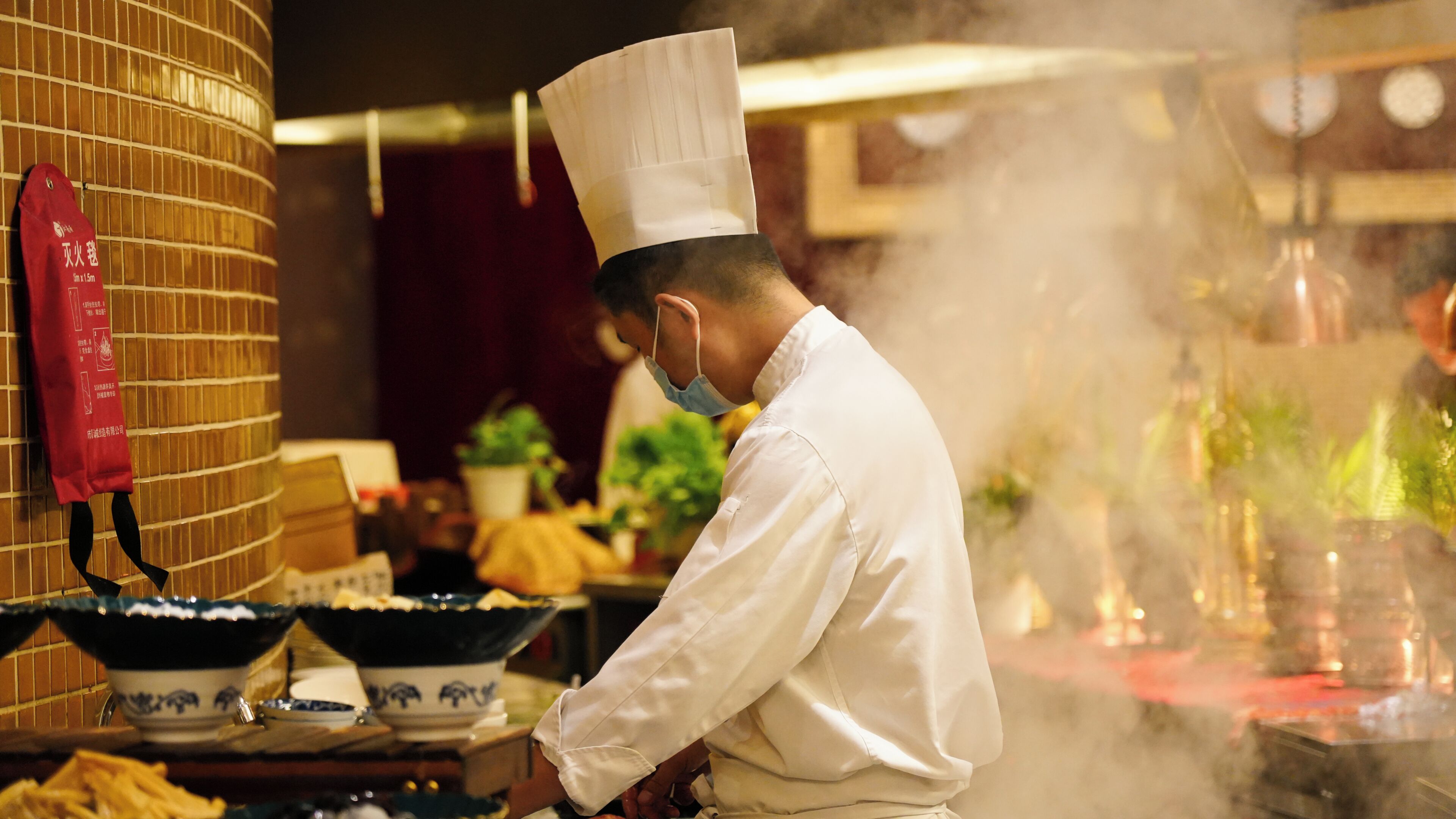 Un chef con uniforme blanco y gorro alto trabajando en una cocina con vapor, rodeado de utensilios y una pared de azulejos.