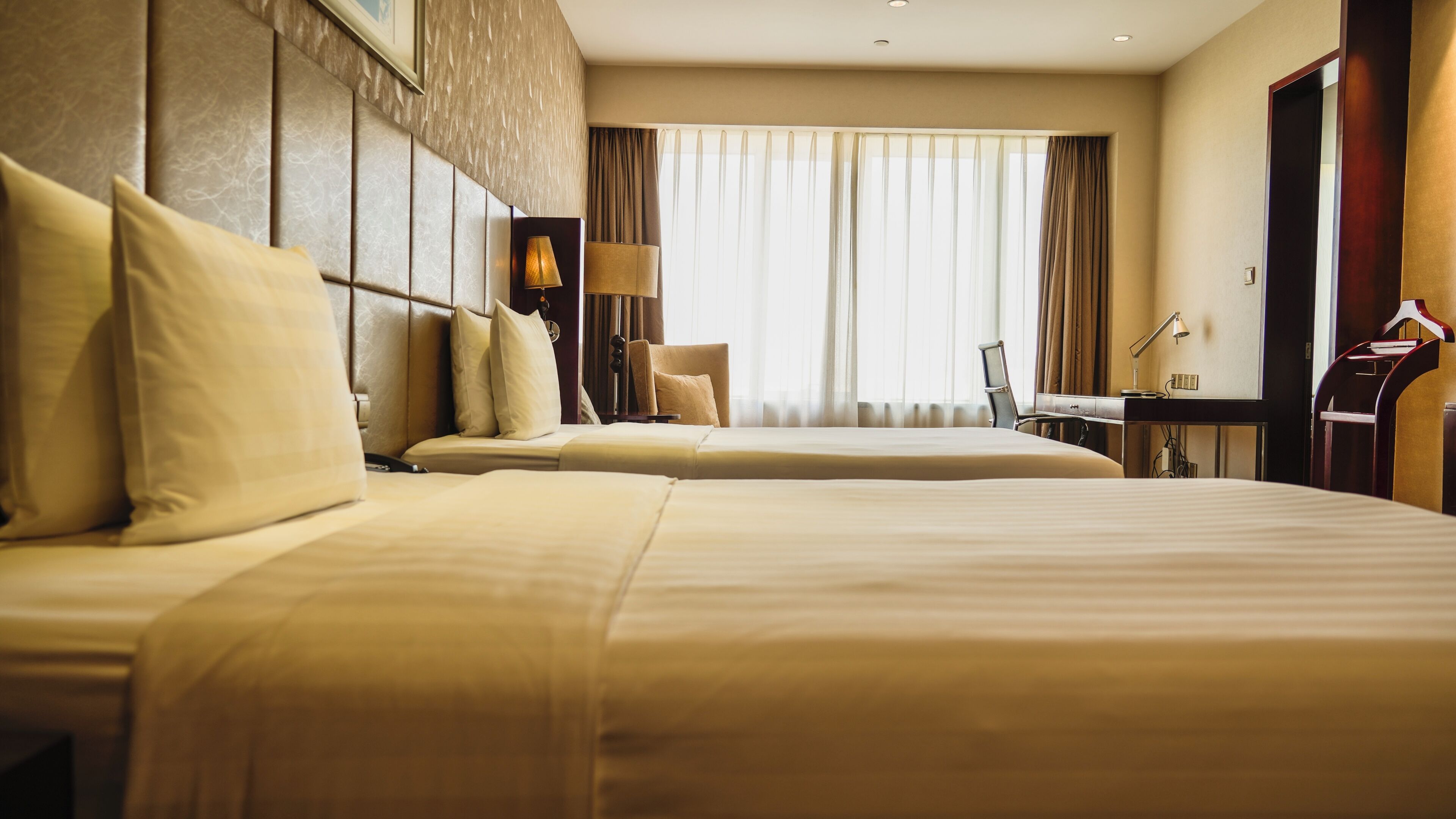 A hotel room with two beds, a desk, a chair, and a large window with curtains.