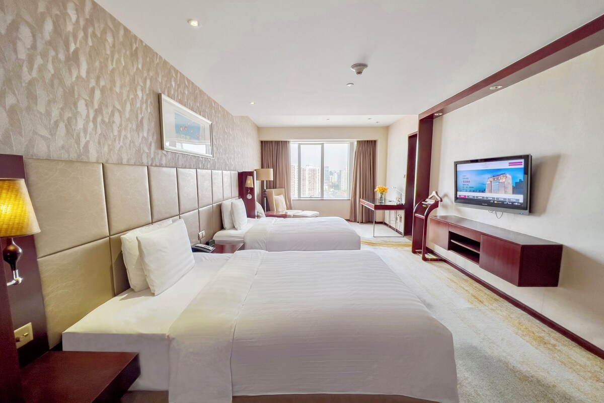 A hotel room with two beds, a desk, a TV, and a window with a city view.