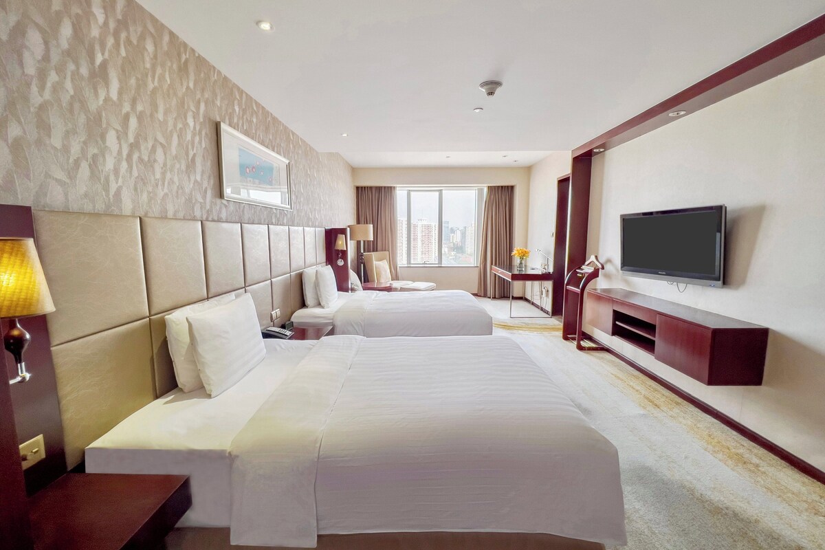 A hotel room with two beds, a TV, a desk, and a window with a city view.