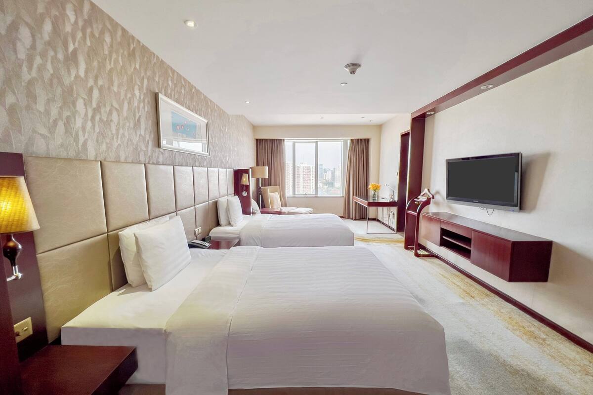 Ein Hotelzimmer mit zwei Betten, einem Fernseher, einem Schreibtisch und einem Fenster mit Blick auf die Stadt.
