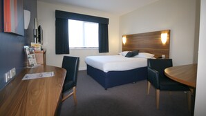 Ein Hotelzimmer mit einem Bett, einem Schreibtisch, zwei Stühlen, einem Holztisch, einem Fenster mit Vorhängen, einer Wandlampe und einem an der Wand befestigten Fernseher.