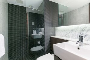 Casa de banho moderna com lavatório branco, chuveiro com azulejos escuros e bancada de mármore.