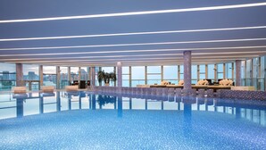 A large indoor swimming pool with clear blue water, surrounded by lounge areas with beige cushions and a cityscape view through large windows. - InterContinental Beijing Beichen by IHG (Beijing)