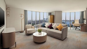 Habitación de hotel moderna con un ventanal amplio que ofrece vistas a la ciudad, un sofá con cojines de colores, una mesa de centro redonda con un florero y un escritorio con una silla.