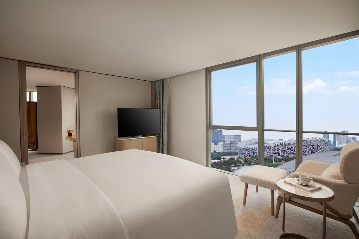 A modern hotel room with a large bed, a flat-screen TV, a small table with a cup and book, and a view of a cityscape through large windows.