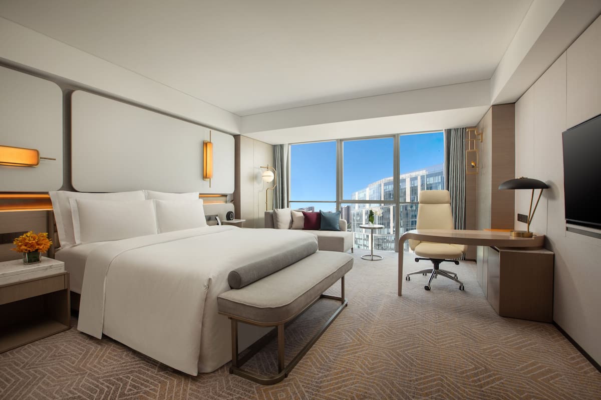 A modern hotel room with a large bed, a desk, a chair, a TV, and a view of the city.