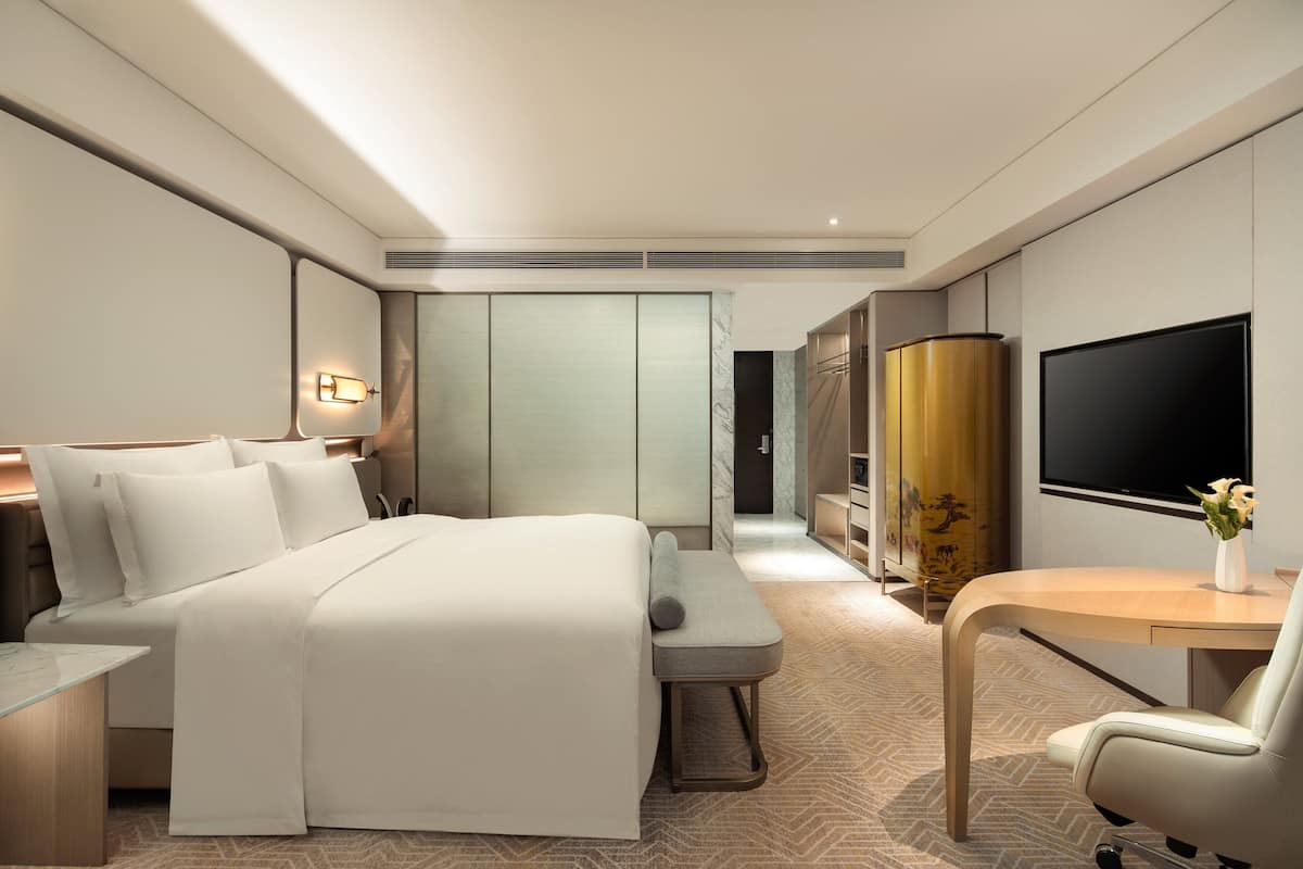A modern hotel room with a large bed, bedside tables, a desk with a chair, a TV, and a wardrobe.