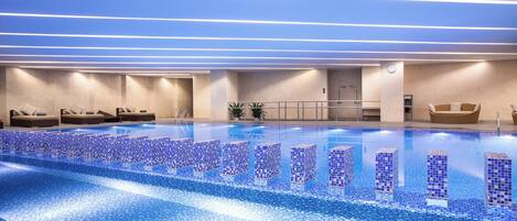 Piscina interior moderna com azulejos de mosaico azul, espreguiçadeiras e ambiente limpo e bem iluminado.