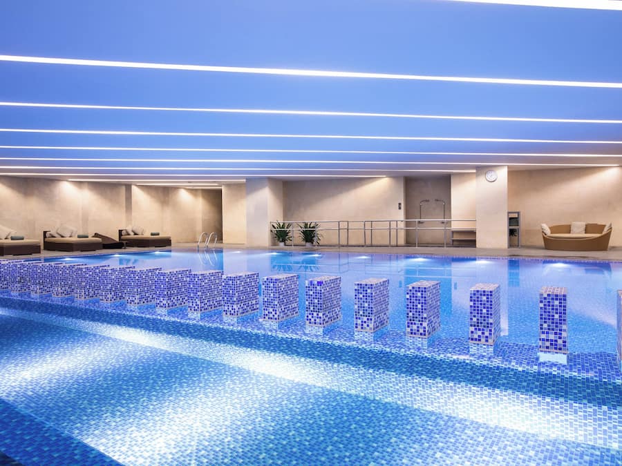 A modern indoor swimming pool with blue mosaic tiles, lounge chairs, and a clean, well-lit environment.