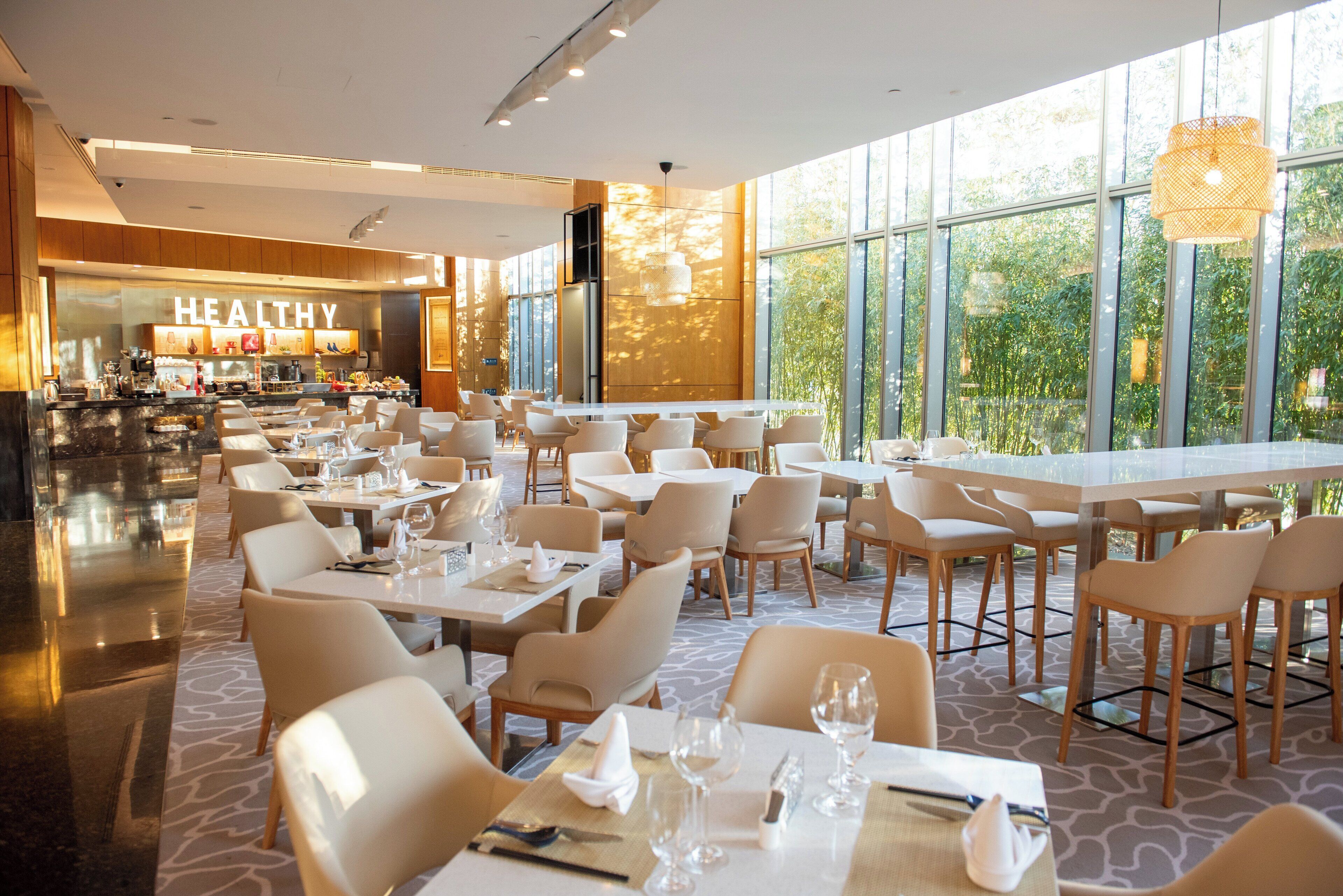 Un restaurante moderno con mesas y sillas blancas, ventanas amplias y un letrero con la palabra 'HEALTHY'.