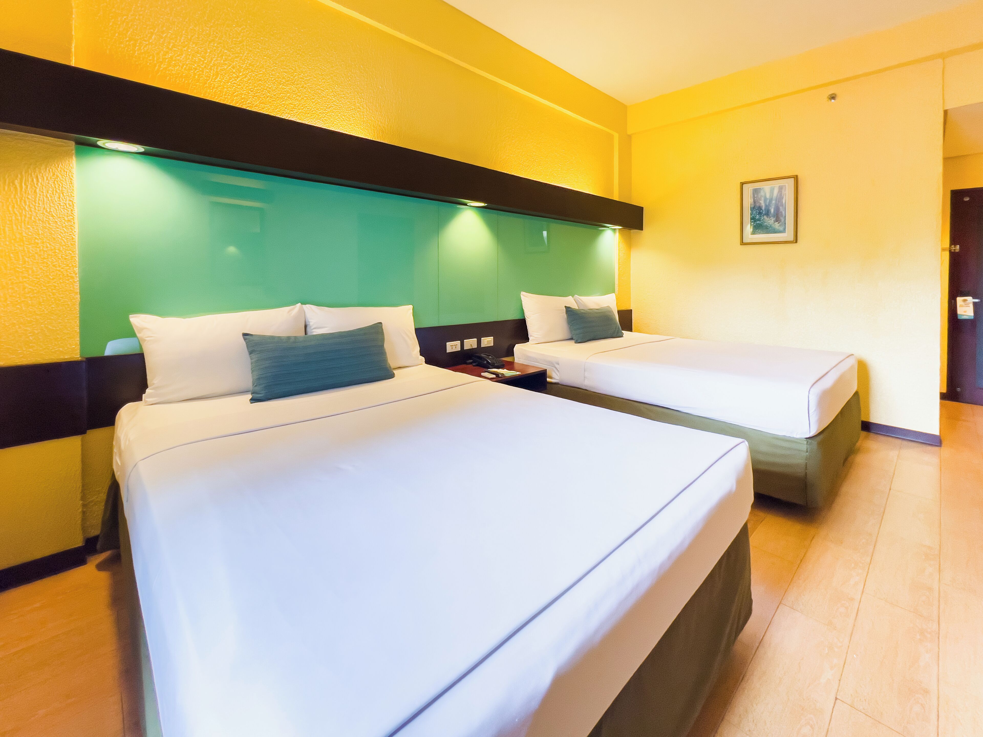Quarto de hotel com duas camas, cada uma com lençóis brancos e uma única almofada. O quarto possui uma parede de destaque verde e uma cabeceira preta.