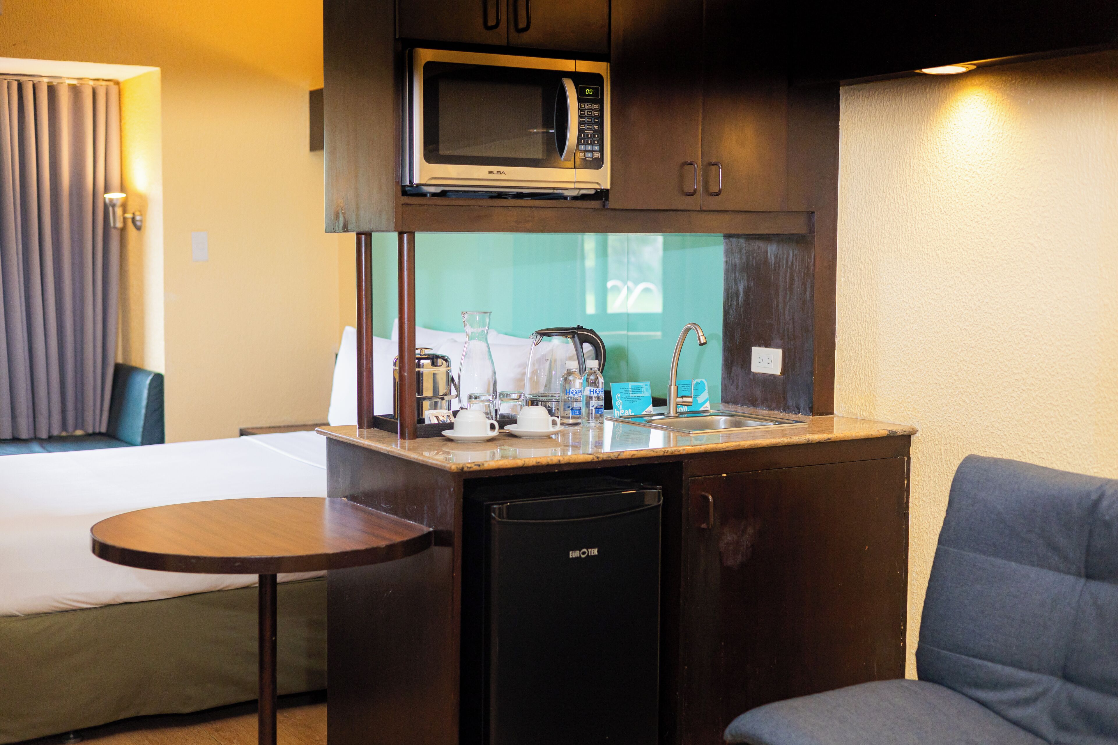 A compact kitchenette with a microwave, sink, and small refrigerator. There is a bed and a chair in the room.