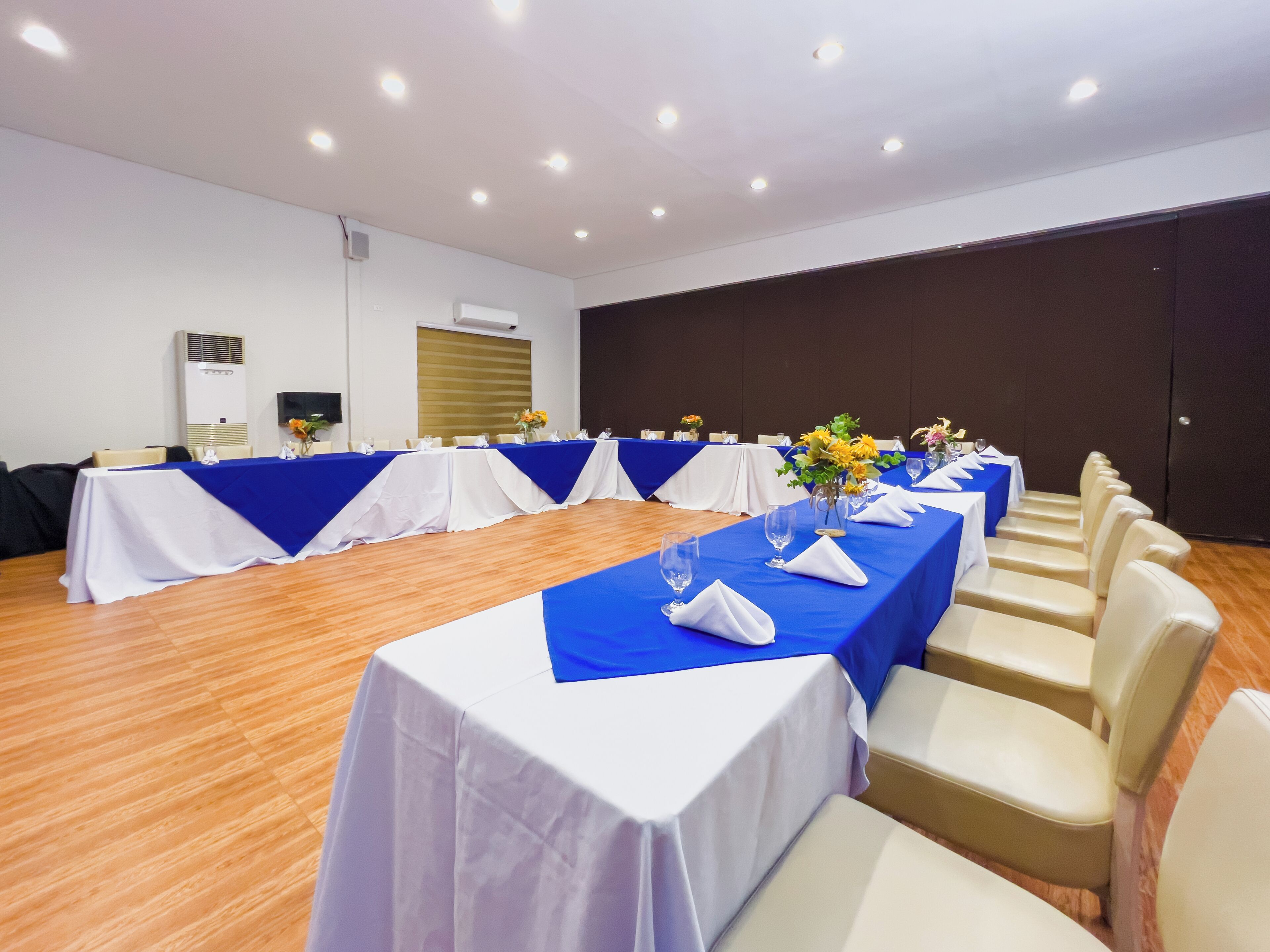 A conference room with multiple tables, each covered with a blue tablecloth and white table runners. The room has wooden flooring and is equipped with ceiling lights.